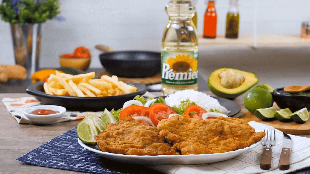 Vista de un plato de chuleta de cerdo apanada, al fondo se ve un frasco de aceite Premier, aguacate y papas.