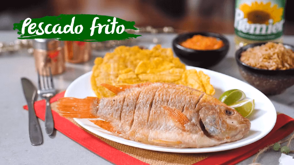 Pescado frito acompañado de tostadas, rodajas de limón en un plato blanco, sobre un individual. Alrededor hay cubiertos, guisos y Aceite Premier.