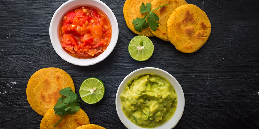 Vista superior de varias arepas de huevo, acompañadas de recipientes con salsas y limones. Todo sobre un fondo negro.