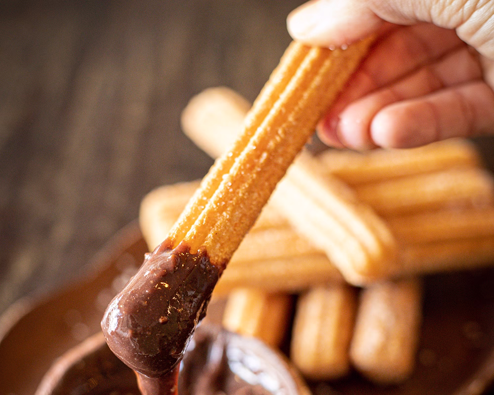 Vista de una mano untando un churro en salsa de chocolate en un bowl pequeño. Al fondo se ven más churros