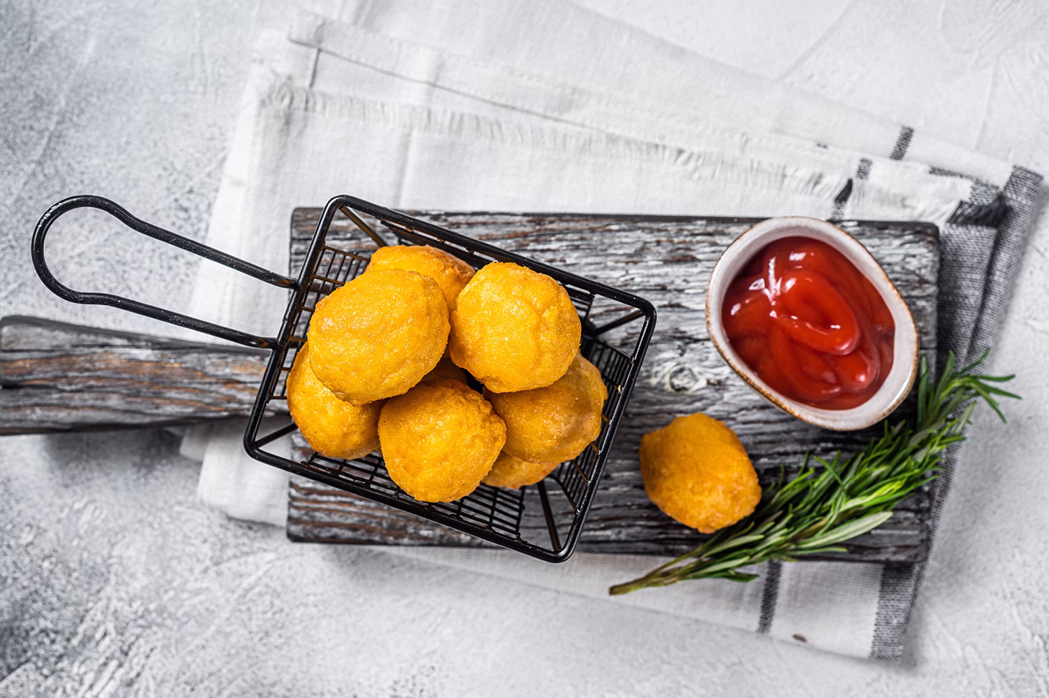 Vista superior de varias papas rellenas en una canasta negra, acompañada de una salsa roja, sobre una tabla de madera. Todo sobre un fondo de mármol.