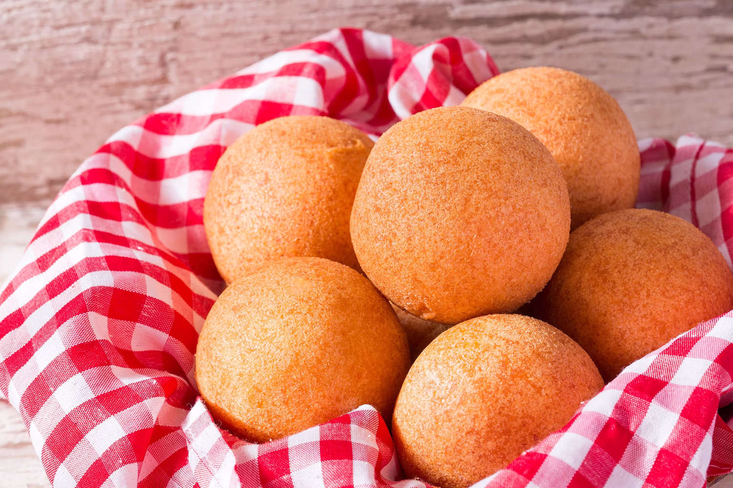 Vista de buñuelos en una canasta decorada con un servilleta de tela a cuadros rojos y blancos. Al fondo se aprecia una superficie de madera.