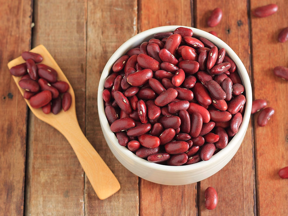 Recipiente blanco con fríjoles rojos, acompañados de una cuchara de madera con más granos al fondo sobre una superficie de madera natural.